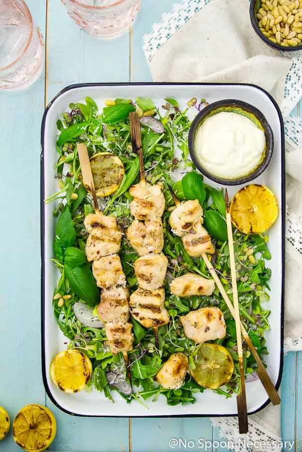 Overhead shot of a tray of Grilled Honey Lemon Chicken Skewers on a bed of Arugula and Basil Salad with a neutral linen, pink water glasses, grilled lemon halves and a bowl of pine nuts framing the photo