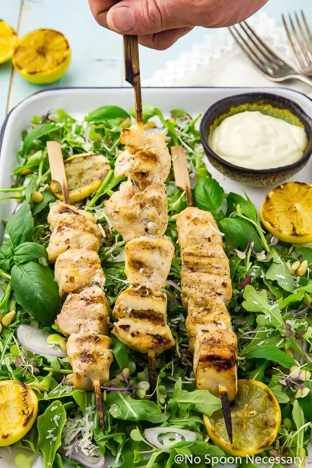 A hand grabbing a chicken skewer from a tray of Grilled Honey Lemon Chicken Skewers on a bed of Arugula and Basil Salad