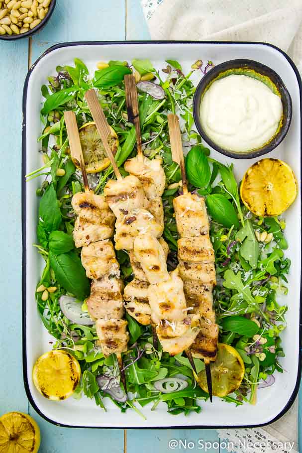 Overhead shot of a tray of Grilled Honey Lemon Chicken Skewers on a bed of Arugula and Basil Salad; with a neutral linen, a bowl of pine nuts and lemon halves framing the photo