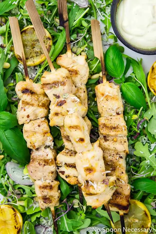 Overhead up-close shot of four overlapping Grilled Honey Lemon Chicken Skewers on a bed of Arugula and Basil Salad, with a bowl of lemon yogurt dip in the right corner