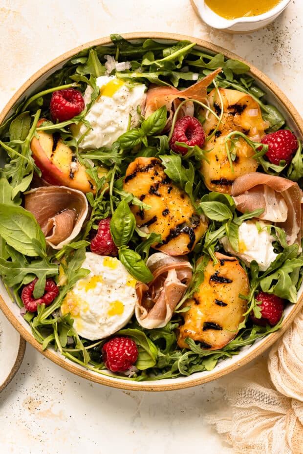 Overhead photo of burrata and peach salad in a large serving bowl with a jar of vinaigrette next to the bowl.