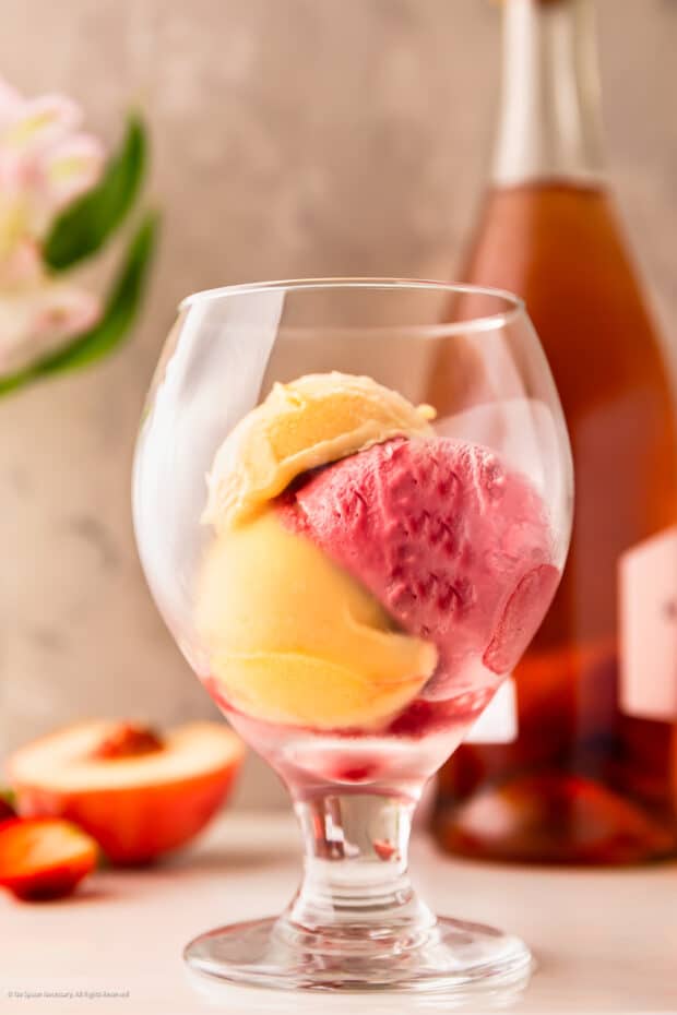 Straight on photo of three scoops of sorbet in a stemmed glass with a bottle of rose in the background.