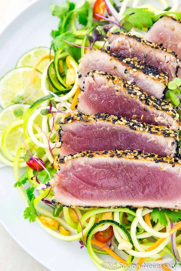Overhead, up-close shot of rare, pan seared, spicy sesame crusted ahi tuna slices over a bed of Thai zucchini noodles with lime slices in the background