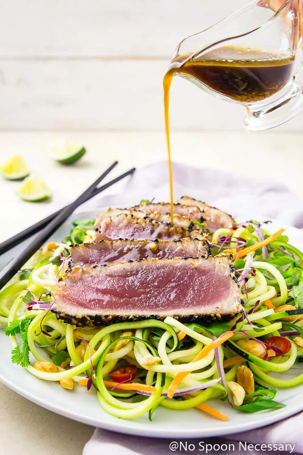 Angled shot of Asian dressing being poured over rare, pan seared, spicy sesame crusted tuna slices on a bed of Thai zucchini noodles with black chopsticks and lime wedges in the background