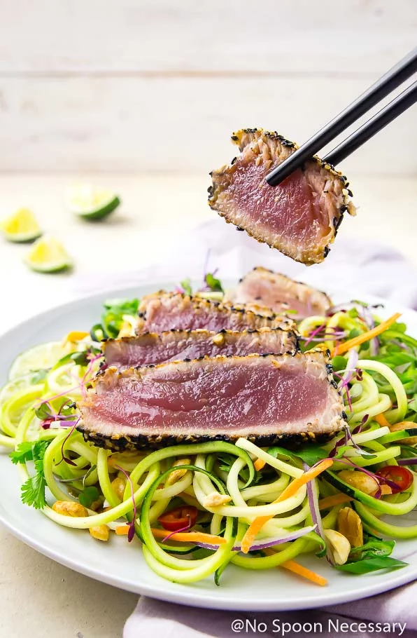 Angled shot of black chopsticks picking up a slice of rare, pan seared, spicy sesame crusted tuna off a white plate of Thai zucchini noodles topped with a sliced filet of sesame crusted tuna