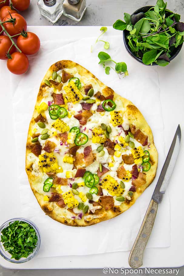Overhead shot of Tex-Mex Charred Corn, Bacon & Jalapeno Pizza on a white square board with vine ripe tomatoes, salt and pepper shakers, knife and ramekins of microgreens and chopped cilantro
