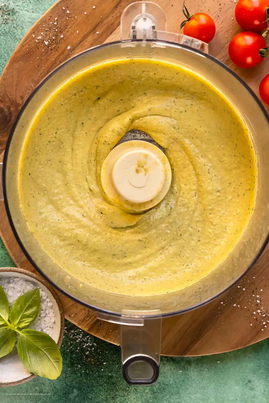 Overhead photo of a creamy green dip in the bowl of a food processor with a bowl fresh basil leaves off to the side.