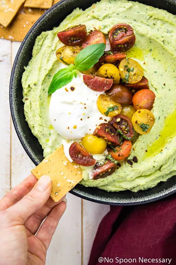 Overhead shot of a pita chip being dipped into a black bowl of basil hummus topped with burrata and caprese tomatoes with a red linen and pile of pita chips on the sides of the bowl