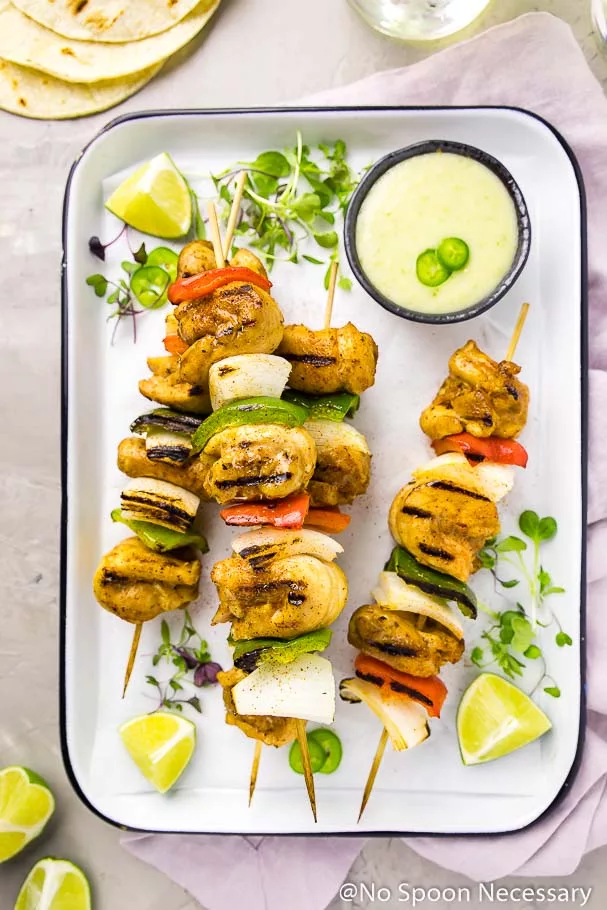 Margarita Fajita Chicken, Onion and Pepper Kebabs on a white tray with micro-greens, lime wedges and a ramekin of pineapple aioli with a purple linen, tortillas and clear glasses surrounding tray