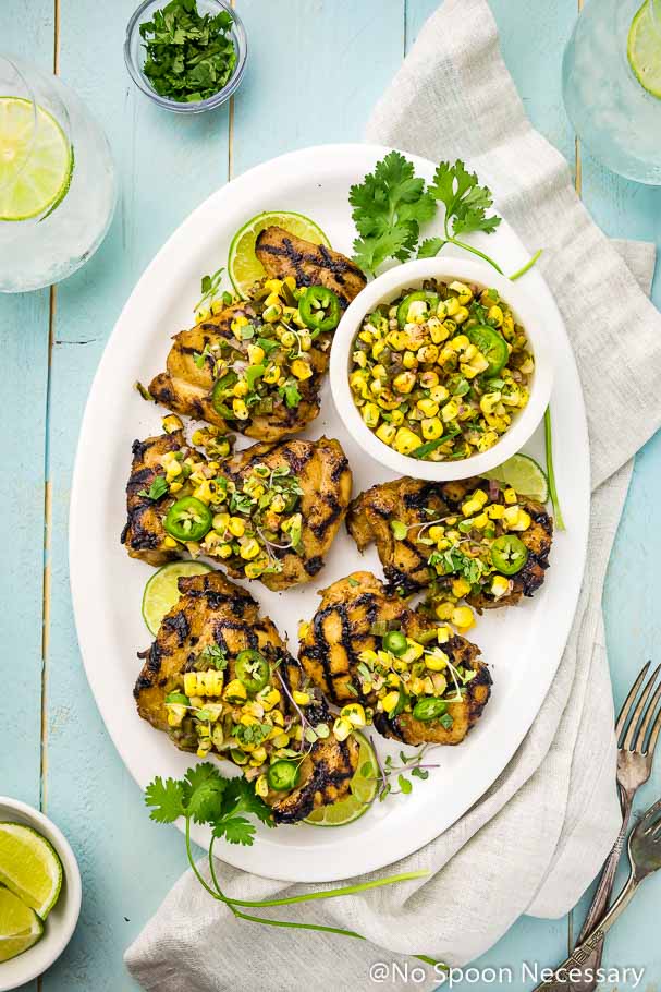 Overhead shot of a white platter of Southwest Buttermilk Grilled Chicken topped with corn salsa on a light blue wood board with glasses of lime water, neutral linen, cilantro, lime wedges and forks surrounding the platter
