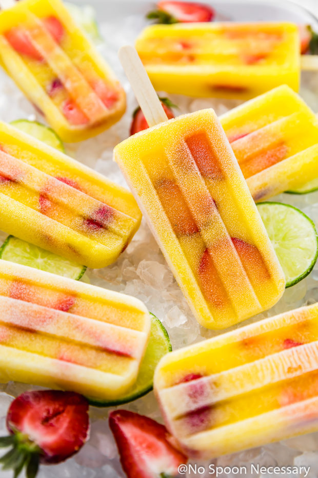 Angled shot of Strawberry Mango Gin and Tonic Boozy Popsicles on a white tray filled with ice with lime slices and strawberries strewn around the popsicles.