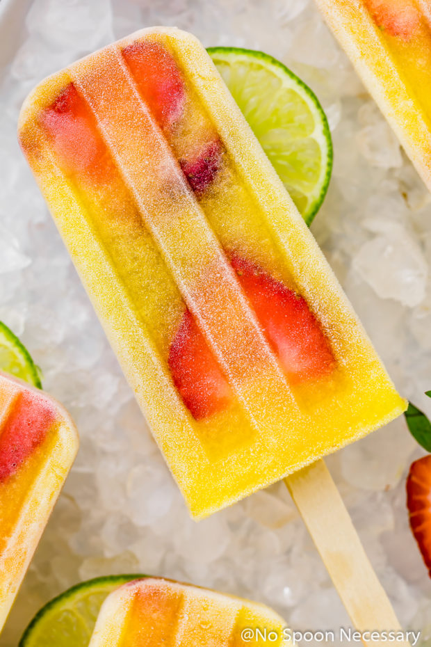 Overhead, up close shot of a Strawberry Mango Gin and Tonic Boozy Popsicle on a surface of ice with more popsicles, lime slices and strawberries peeking in the frame.