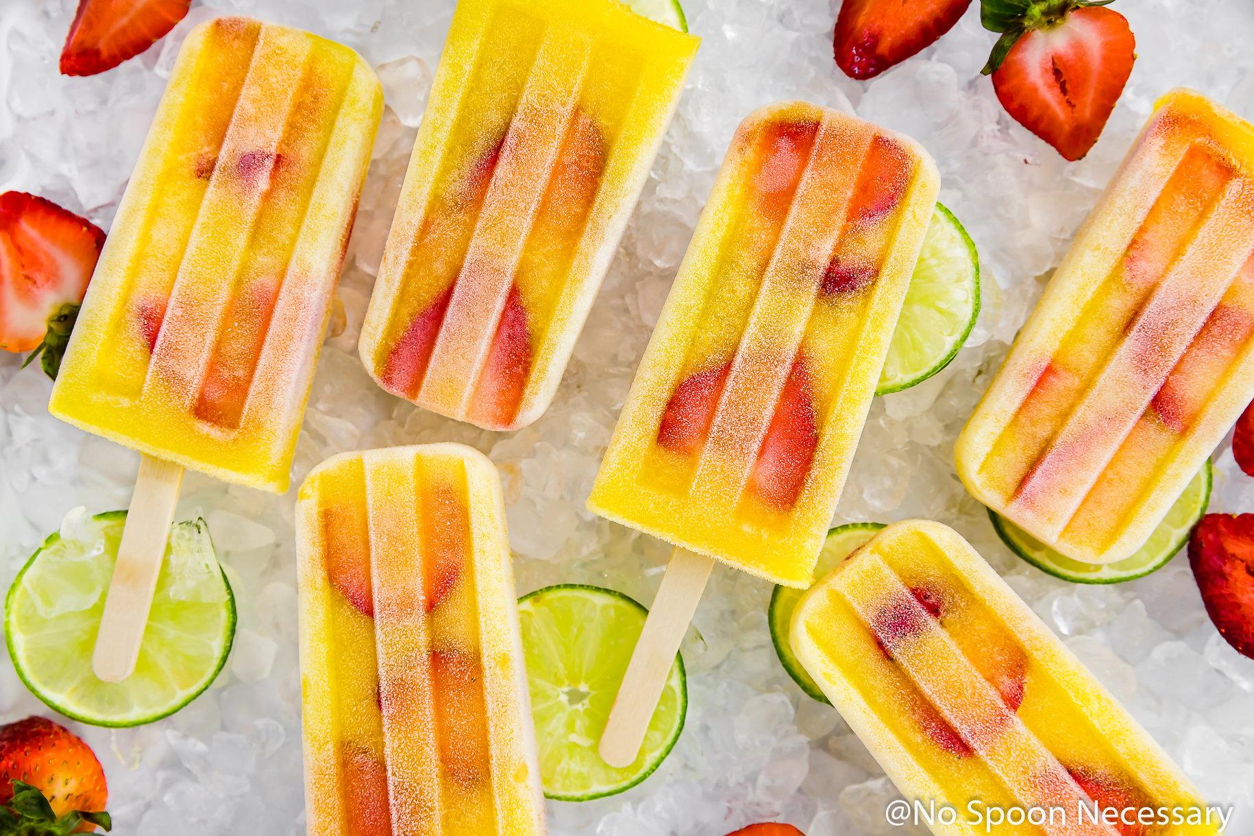Overhead, landscape shot of Strawberry Mango Gin and Tonic Boozy Popsicles on a surface of crushed ice with lime slices and strawberries strewn around the popsicles.