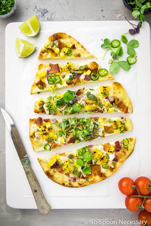 Overhead shot of a sliced Tex-Mex Charred Corn, Bacon & Jalapeno Pizza on a white board with lime wedges, knife, vine ripe tomatoes and ramekins of microgreens and chopped cilantro in the corners