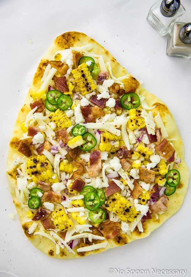 Overhead shot of an unbaked Tex-Mex Charred Corn, Bacon & Jalapeno Pizza on a sheet of parchment paper with small salt and pepper shakers in the upper corner