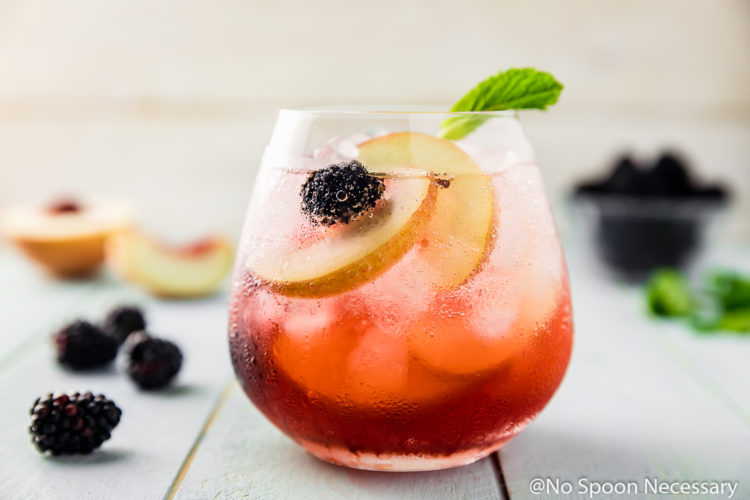 Straight on shot of a Blackberry Peach Elderflower Cocktail garnished with fresh blackberries, peach slices and mint with more fresh fruit surrounding the cocktail glass.