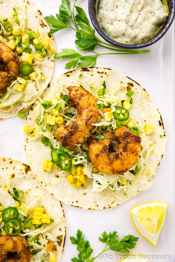 Overhead shot of an open Cajun Shrimp Taco on a white board with 2 other tacos, fresh parsley, lemon wedges and a ramekin of remoulade surrounding it.