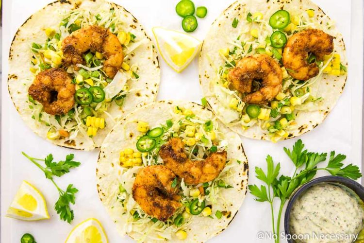 Overhead, landscape shot of three Cajun Shrimp Tacos with Spicy Corn Slaw & Remoulade on a white board with lemon wedges, slices of jalapeños, fresh parsley and a small ramekin of remoulade.