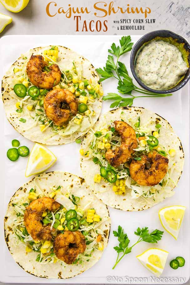Overhead shot of three Cajun Shrimp Tacos with Spicy Corn Slaw & Remoulade on a white board with lemon wedges, slices of jalapeños, fresh parsley and a small ramekin of remoulade.