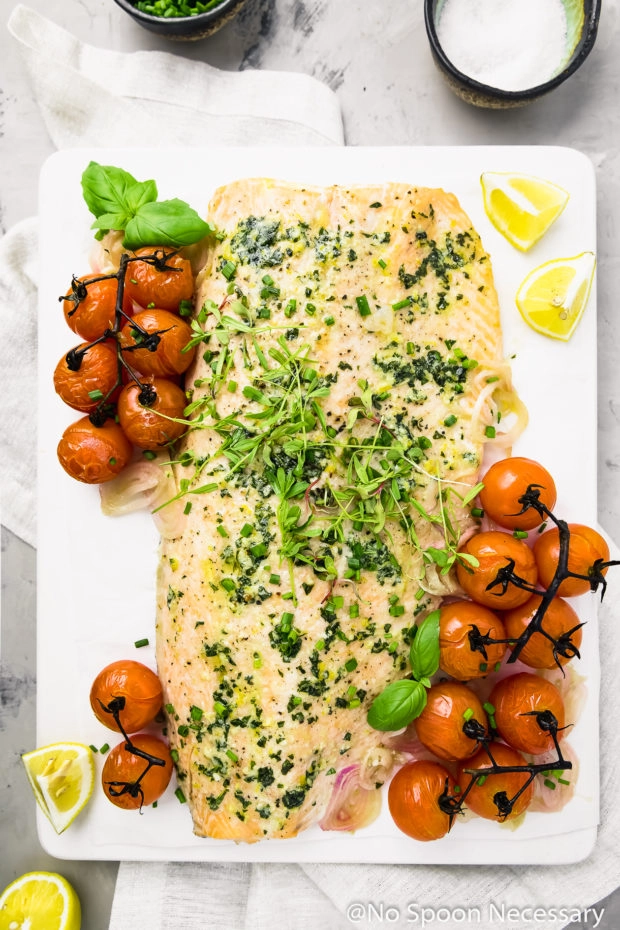 Overhead shot of Oven Baked Basil Butter Salmon with Burst Tomatoes on a white board with a neutral linen, lemon wedges and bowls of chives and sea salt surrounding the board.