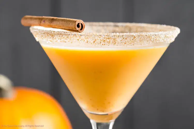 Straight on photo of a pumpkin martini in a martini glass with a cinnamon sugar rim and cinnamon stick.
