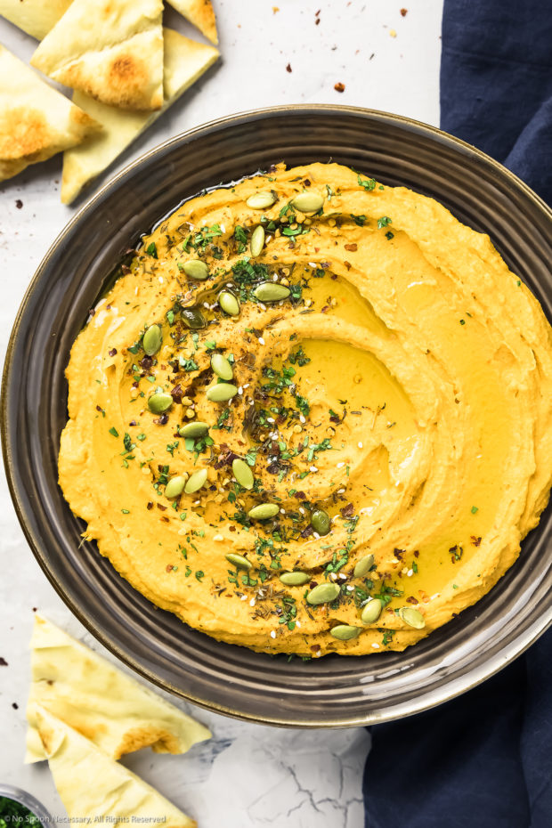 Overhead photo of sweet potato hummus garnished with pumpkin seeds, za'atar and fresh parsley in a large dark brown serving bowl with pita wedges and a navy linen napkin next to the bowl.