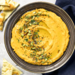 Overhead, landscape photo of sweet potato hummus garnished with pumpkin seeds, za'atar and fresh parsley in a large dark brown serving bowl with pita wedges and a navy linen napkin next to the bowl.