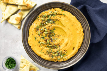Overhead, landscape photo of sweet potato hummus garnished with pumpkin seeds, za'atar and fresh parsley in a large dark brown serving bowl with pita wedges and a navy linen napkin next to the bowl.