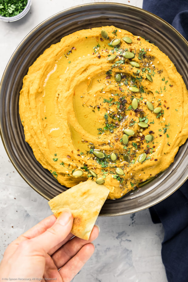 Overhead action photo of Roasted Sweet Potato Hummus in a brown bowl with a hand holding a pita wedge and dipping it into the hummus.