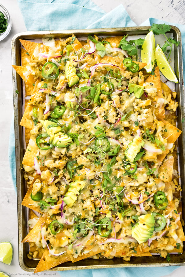 Overhead shot of Wonton Chicken Nachos drizzled with Spicy Peanut Sauce on a small sheet pan with a teal linen underneath the pan and slices of limes and a ramekin of scallions surrounding the pan.
