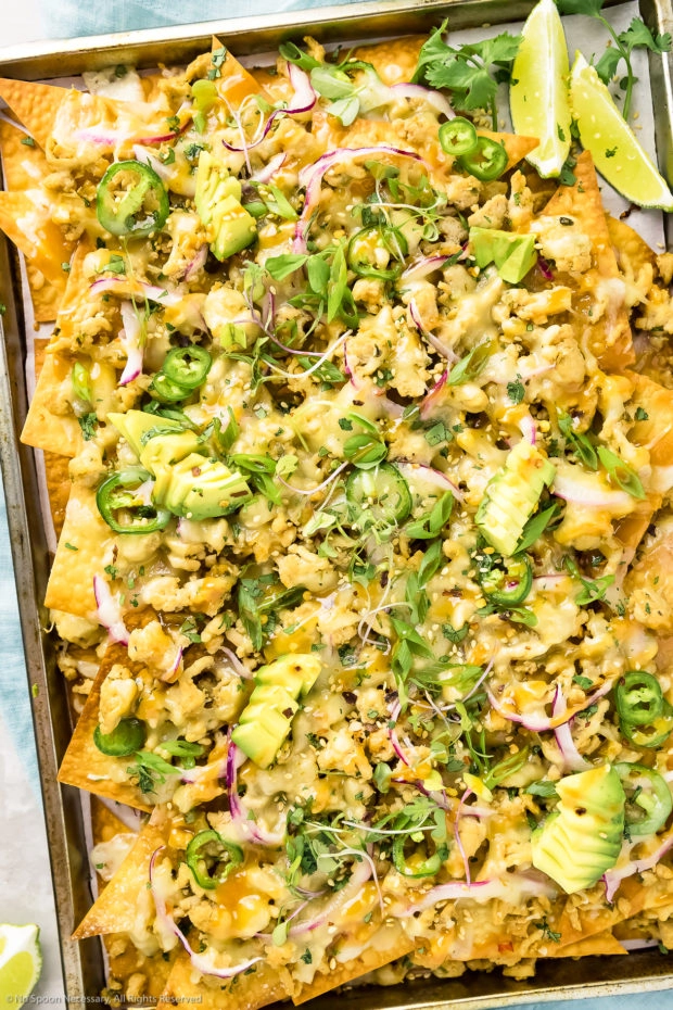 Overhead shot of Chicken Wonton Nachos drizzled with Spicy Peanut Sauce on a small sheet pan with limes and fresh cilantro next to the nachos.