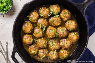 Overhead shot of a cast iron skillet filled with Baked Chicken Apple Meatballs with a blue linen tied around the handle and a ramekin of sliced scallions surrounding the skillet.