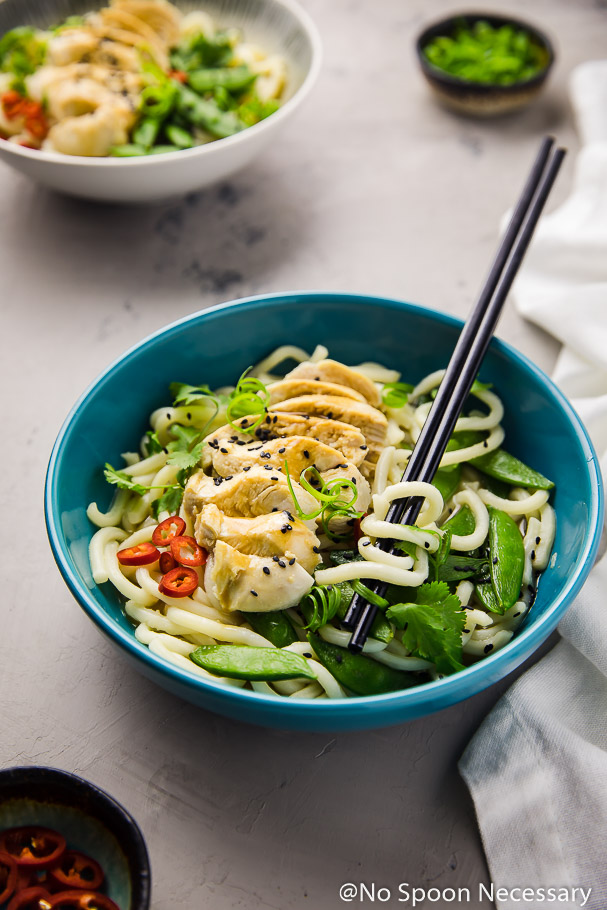 Angled shot of a turquoise bowl filled with Ginger Miso Chicken with Udon Noodles and Snap Peas with black chopsticks intertwined with the noodles; and a small white bowl of Ginger Miso Poached Chicken Noodles, ramekin or scallions, ramekin of red chilies and white linen surrounding the bowl