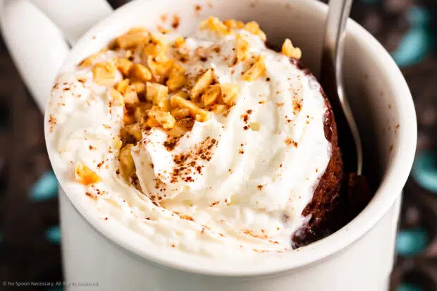 Close-up photo of a Mug Lava Cake topped with whipped cream and crushed nuts in a white mug with a spoon inserted into the mug.