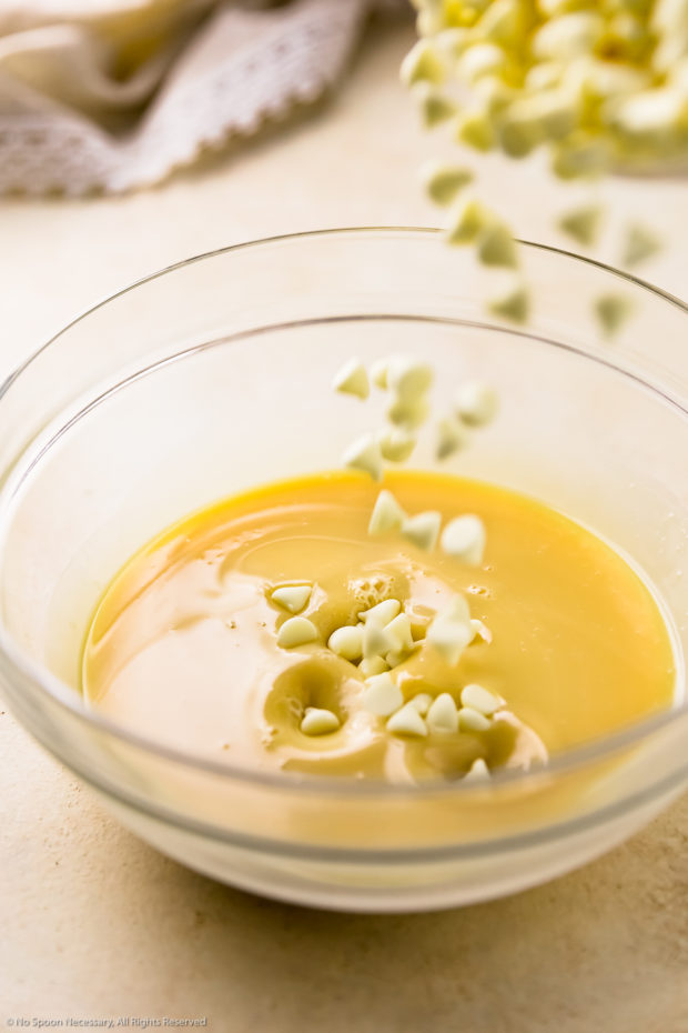 Angled phot of white chocolate chips being poured into a glass bowl containing condensed milk.