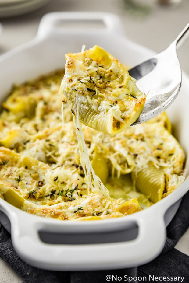 Angled shot of Baked Pumpkin Stuffed Shells topped with Parmesan in a white baking dish with a silver serving spoon lifting up a shell with the melted cheese pulling from the rest of the shells in the baking dish.
