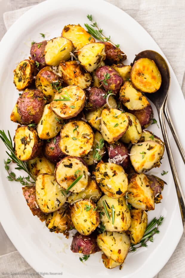 Overhead photo of little roasted potatoes with herbs on a white serving platter with a serving spoon.