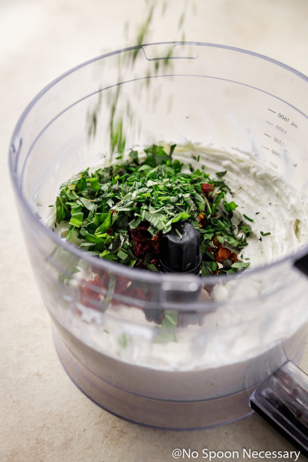 45 degree angle shot of the inside of a food processor bowl containing the ingredients for Baked Sundried Tomato Goat Cheese Dip with fresh herbs being dropped into the bowl