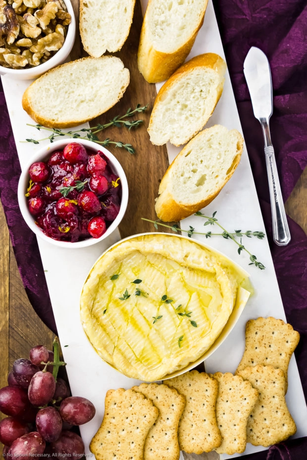 Overhead photo of a white marble cheese board containing Baked Camembert cheese, a bowl of fresh cranberry sauce, slices of baguette, a bowl of walnuts, crackers and red grapes.