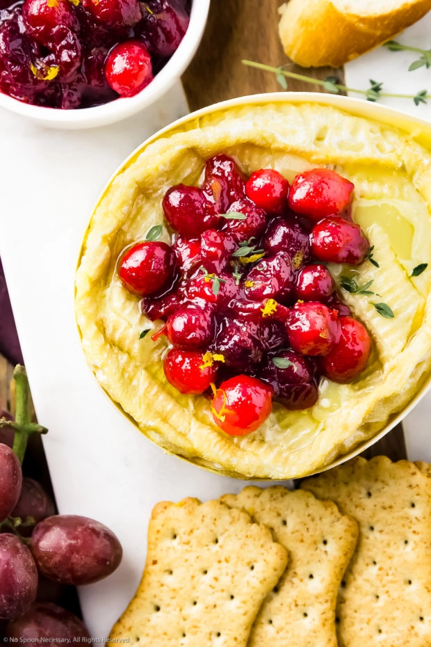 Overhead photo of warm Baked Camembert Cheese topped with fresh Cranberry sauce and surrounded by crackers, red grapes and slices of baguette.