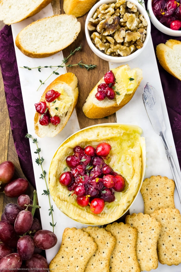 Overhead photo of a white cheese board featuring warm Camembert cheese topped with fresh cranberry sauce, slices of baguettes, a bowl of walnuts, crackers and red grapes, with a dark maroon napkin tucked under the cheese board.