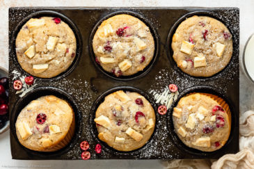 Overhead landscape photo of Chocolate Cranberry Muffins dusted with powdered sugar in a muffin pan with a ramekin of fresh cranberries, glass of milk and pale tan napkin next to the pan.