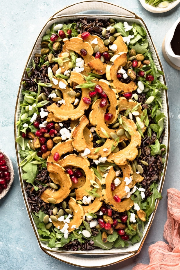 Overhead photo of Wild Rice Salad topped with delicata squash, pomegranate arils and sliced scallions on a white platter with ramekins of pomegranate arils, sliced scallions and a small container of honey balsamic vinaigrette arranged around the salad.