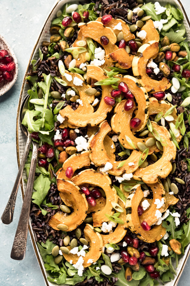 Overhead photo of Wild Rice Salad featuring roasted delicata squash, pomegranate arils and sliced scallions on a white platter with serving spoons inserted into the salad and a ramekin of pomegranate arils next to the platter.