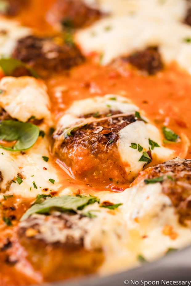 45 degree angle, up-close shot of a spicy baked chicken meatball in vodka sauce topped with melted burrata cheese and garnished with fresh basil and chopped parsley.