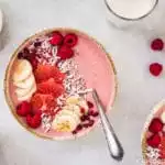 Overhead shot of a Berry Citrus Smoothie Bowl topped with slices of bananas, grapefruit, whole raspberries, pomegranate arils and shredded coconut. There is another smoothie bowl, 2 milk glasses, and ramekins of honey and fresh raspberries surrounding the bowl.