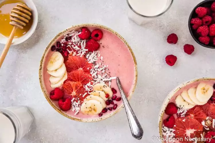 Overhead shot of a Berry Citrus Smoothie Bowl topped with slices of bananas, grapefruit, whole raspberries, pomegranate arils and shredded coconut. There is another smoothie bowl, 2 milk glasses, and ramekins of honey and fresh raspberries surrounding the bowl.