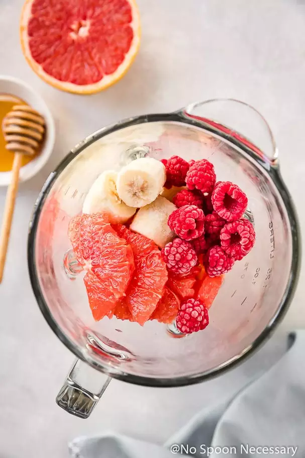 Overhead shot of a blender filled with fresh, frozen fruit (grapefruit, bananas and raspberries) to make a Berry Citrus Smoothie Bowl. Blender is surrounded by a ramekin of honey, grapefruit half and pale blue linen.
