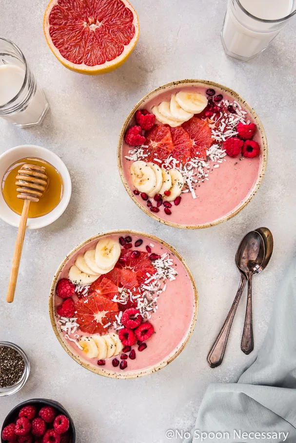 Overhead shot of two Berry Citrus Smoothie Bowls topped with slices of bananas, grapefruit, whole raspberries, pomegranate arils and shredded coconut. The bowls are surrounded by glasses of milk, a grapefruit half, 2 spoons, pale blue linen and ramekins of honey, chia seeds and raspberries.
