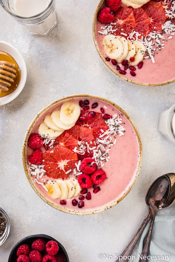 Overhead shot of a Berry Citrus Smoothie Bowl topped with slices of bananas, grapefruit, whole raspberries, pomegranate arils and shredded coconut. The bowl is surrounded by another smoothie bowl, glasses of milk, 2 spoons, pale blue linen and ramekins of honey and raspberries.
